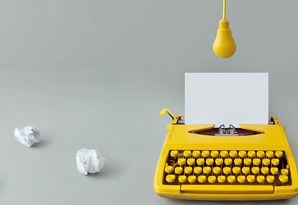 Gelbe Schreibmaschine mit eingespanntem Papier. Darüber eine gelbe Glühbirne. Daneben 3 zerknüllte weiße Papiere ©AdobeStock/Pixelbliss